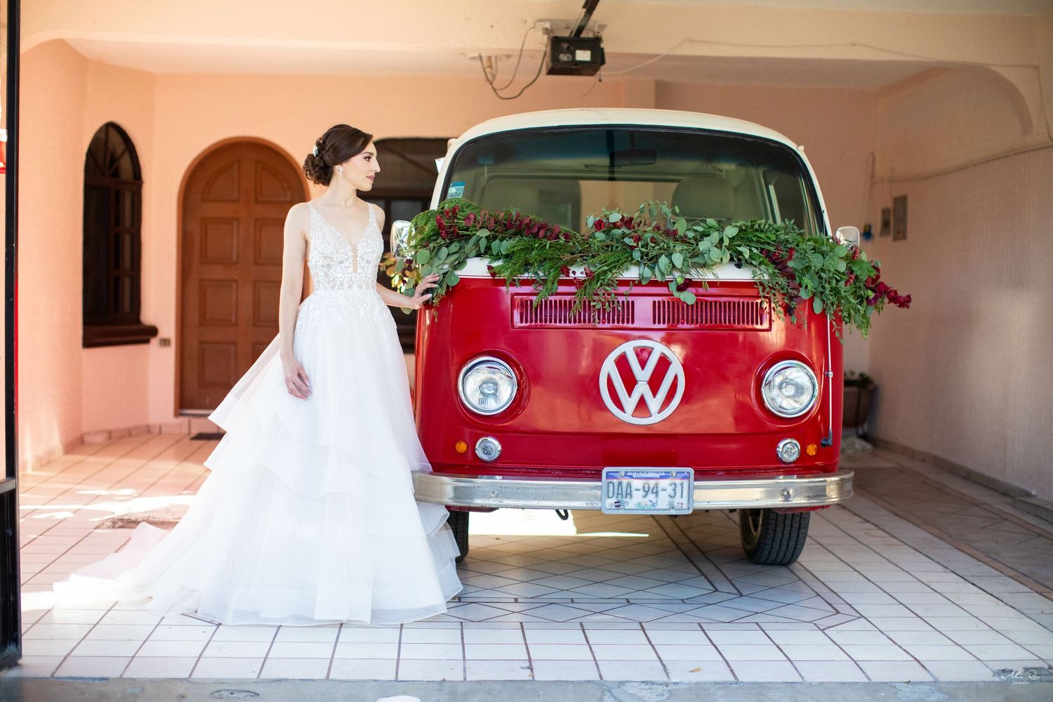 Bride in layered white wedding gown posing beside vintage red Volkswagen bus decorated with flowers.