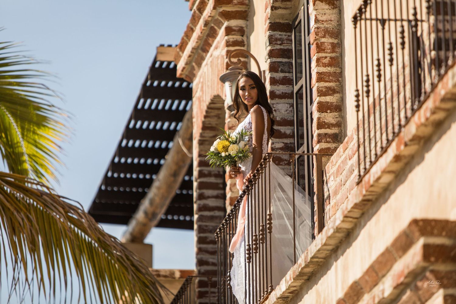 Bride in white gown holding yellow bouquet on brick balcony with palm tree and blue sky.