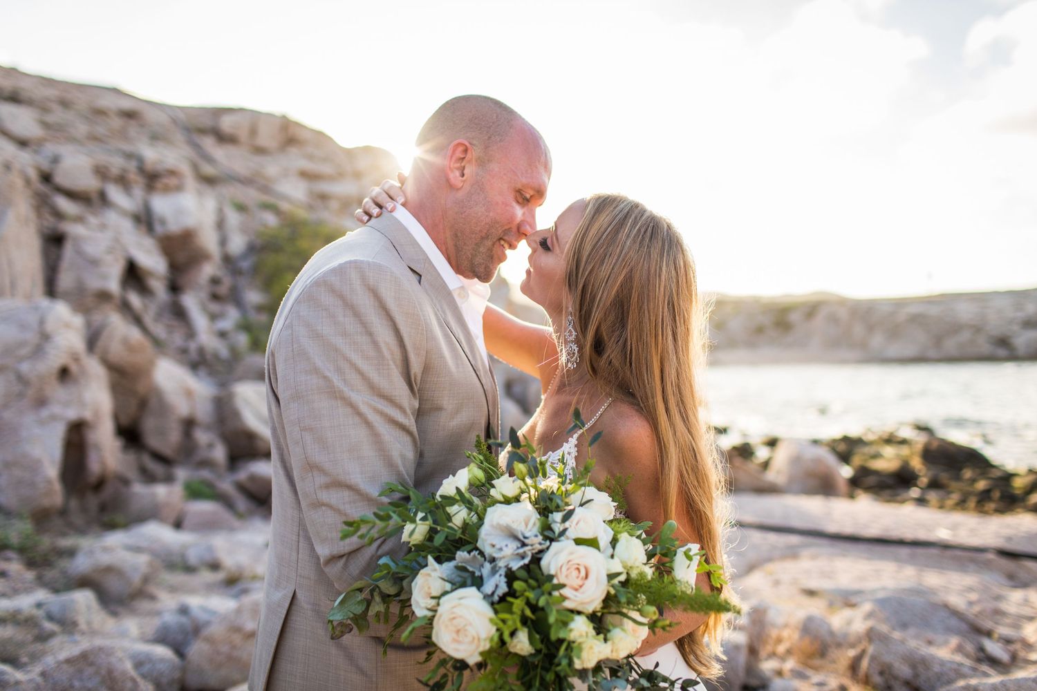 Bride holding white floral bouquet and groom share a tender moment at a rocky beach sunset.