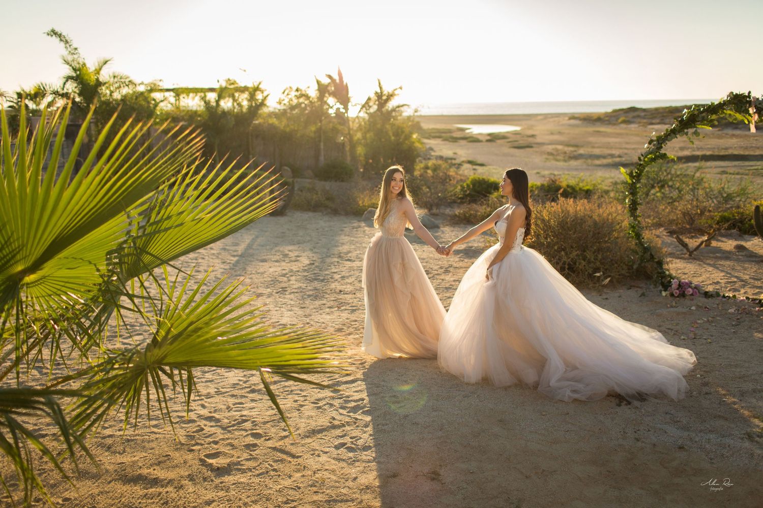 Two brides in tulle gowns hold hands facing each other near a floral arch on a sandy beach at sunset.