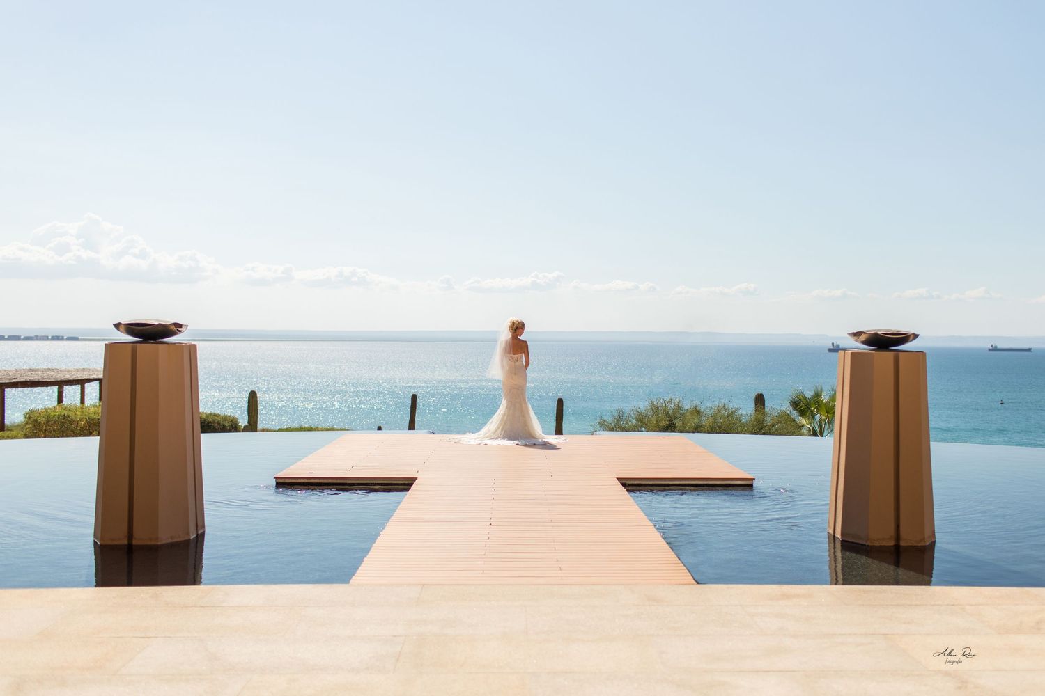 Bride in white gown stands on wooden dock overlooking serene blue ocean at luxury resort wedding venue.