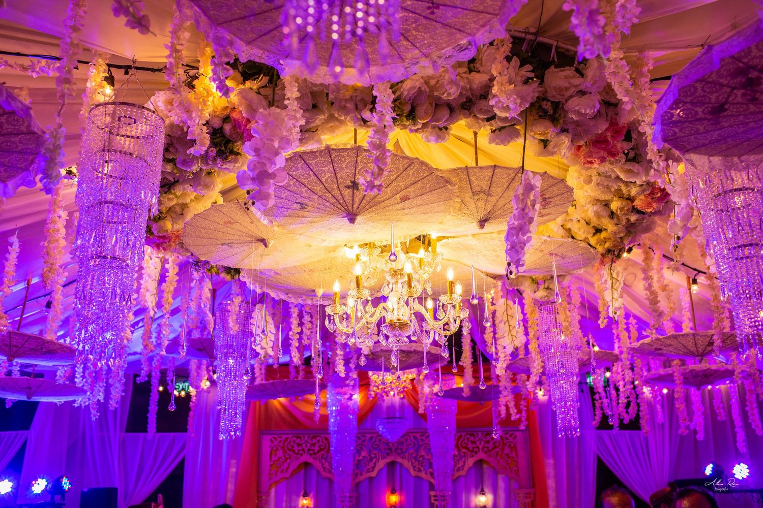 Opulent Indian wedding venue with floral chandeliers, crystal strands, and vibrant purple lighting.