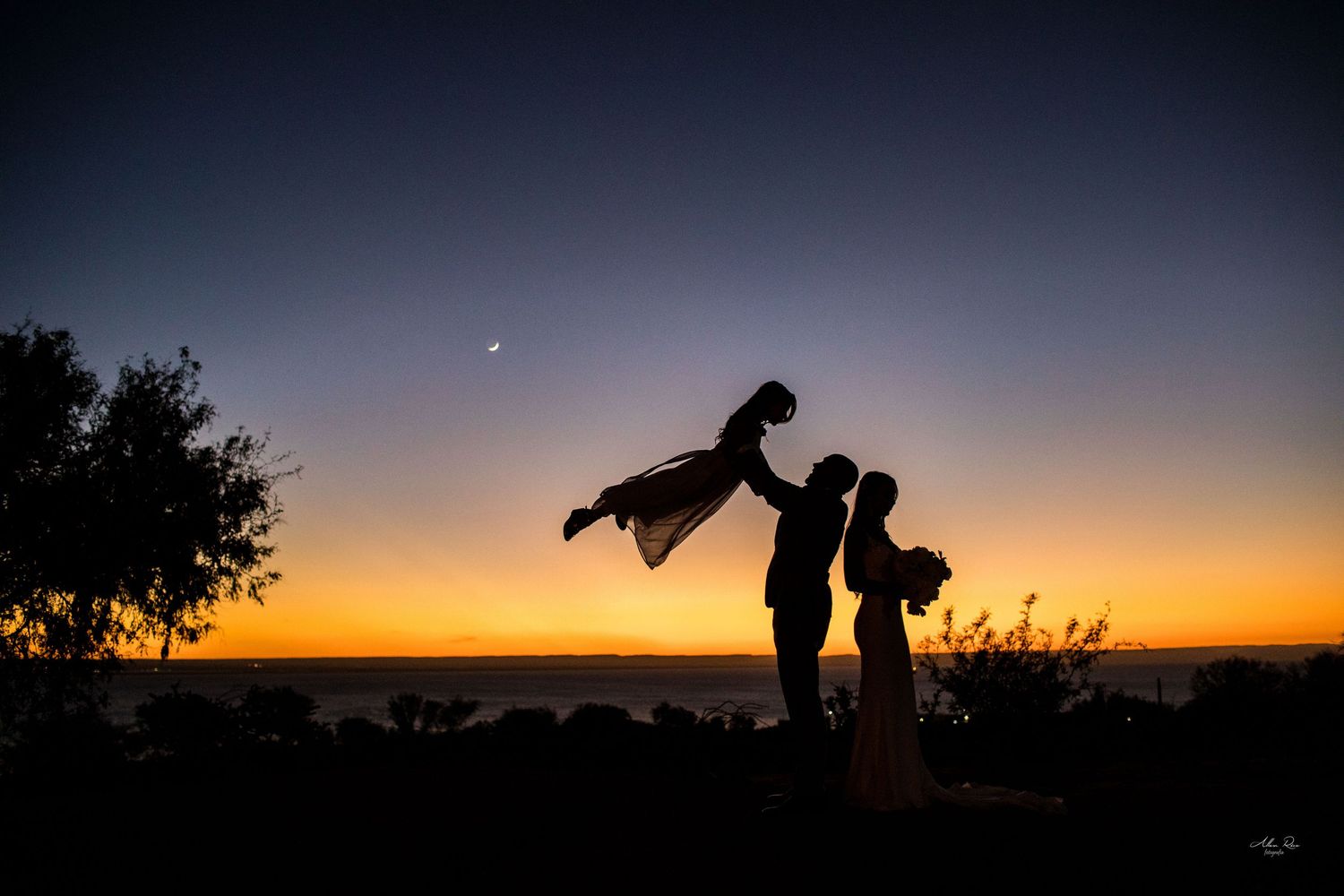 Silhouette of a couple at sunset, groom lifting bride against vibrant orange and blue sky.