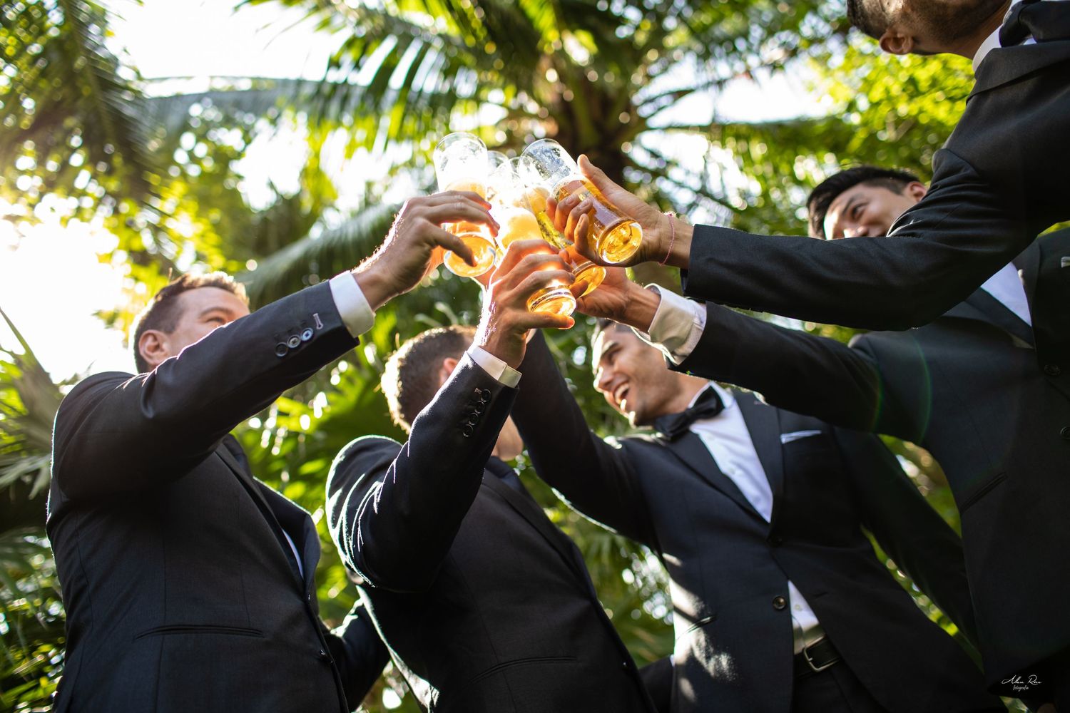 Groomsmen in black suits toast with beer glasses surrounded by lush tropical greenery at wedding celebration.