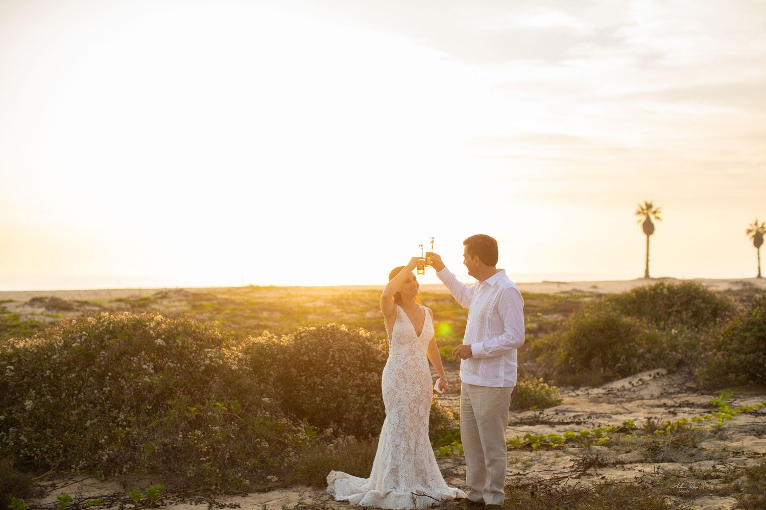 Bride and groom toasting champagne glasses at golden sunset on scenic coastal landscape.