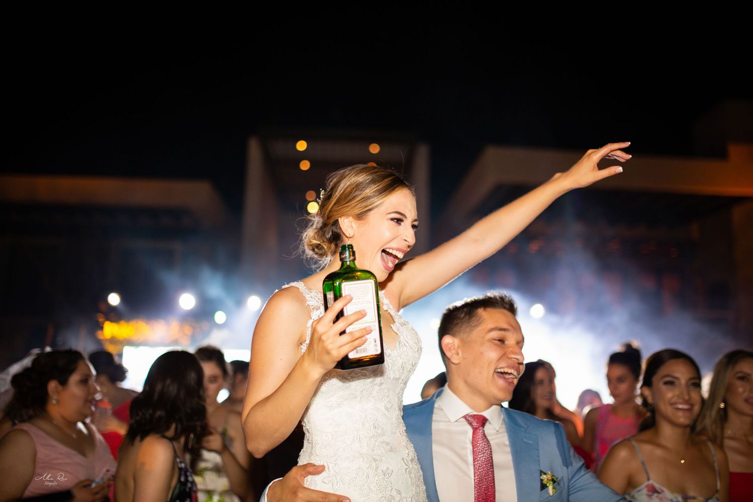 Joyful bride holding bottle aloft on grooms shoulders celebrating at outdoor wedding reception.