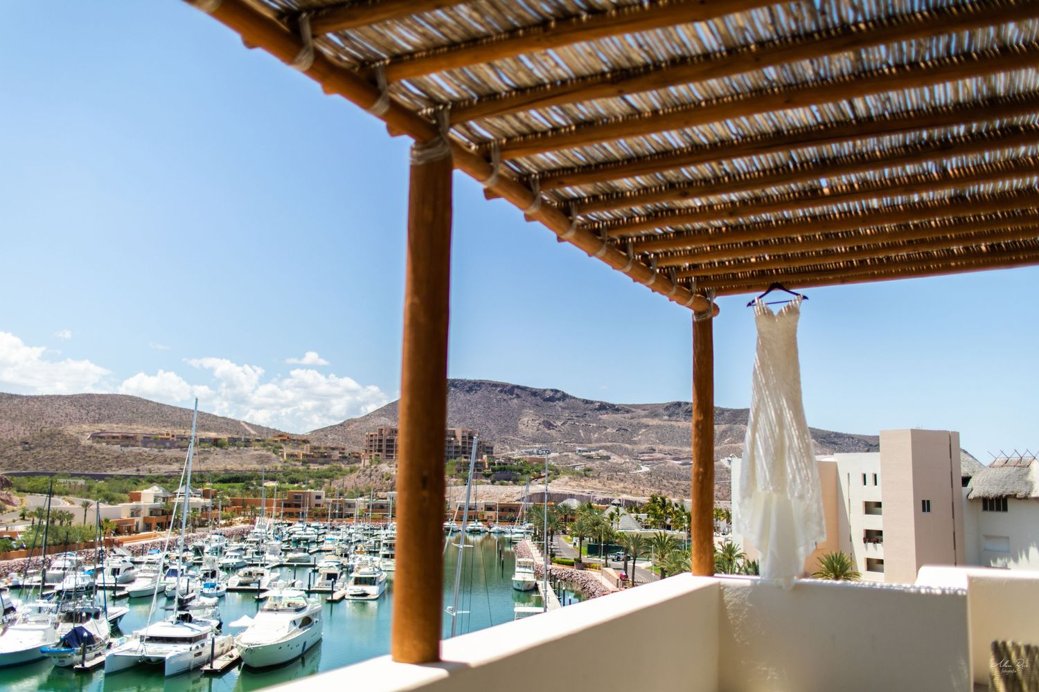 Elegant wedding dress hanging under a rustic pergola overlooking a marina with boats and mountains.