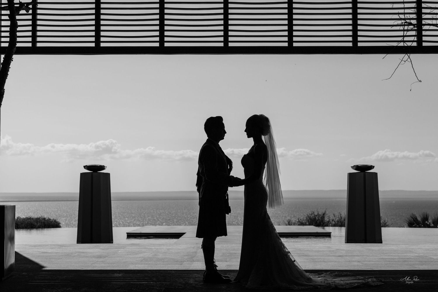 Silhouette of bride and groom holding hands at sunset by the ocean in a romantic black and white wedding photo.