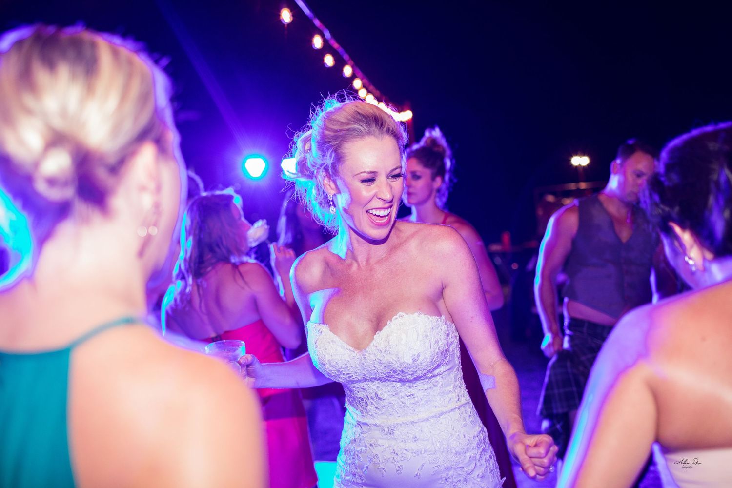 Bride in strapless lace wedding gown dancing joyfully at outdoor wedding reception under blue and purple lights.