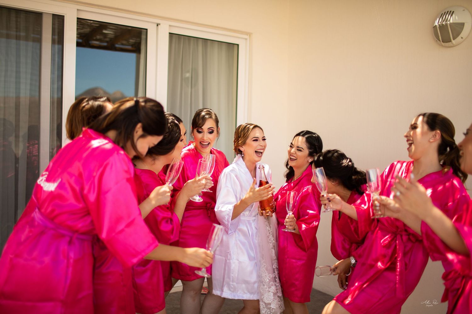Bride in white robe celebrating with bridesmaids in matching pink robes holding champagne glasses.