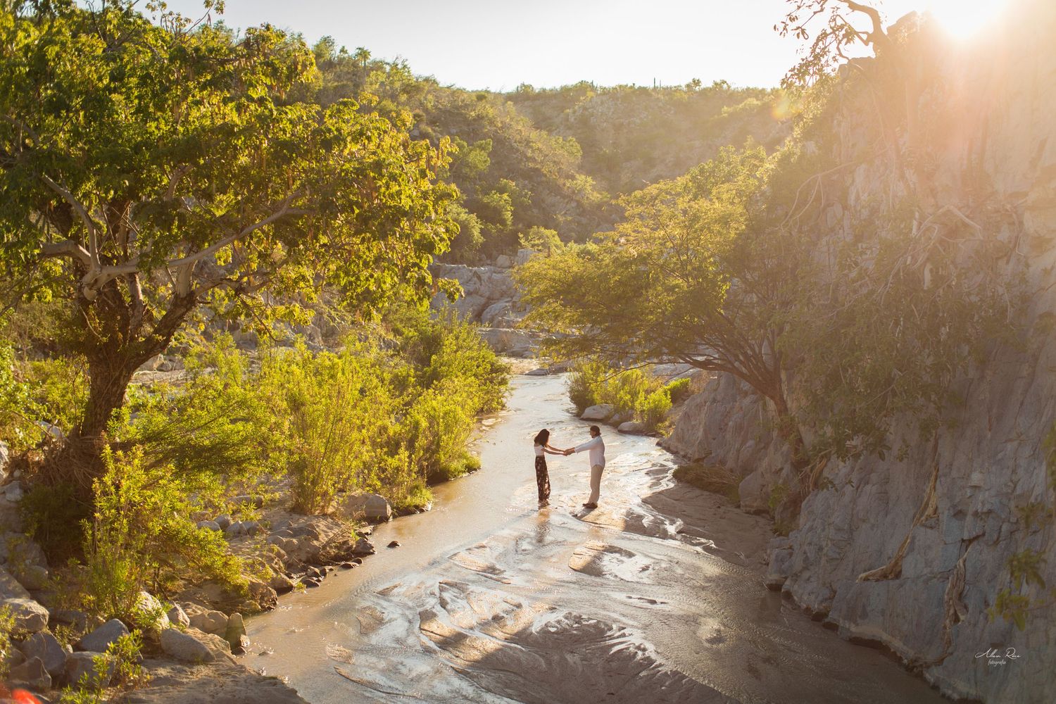 Couple holding hands in a scenic sunlit river canyon surrounded by lush trees.