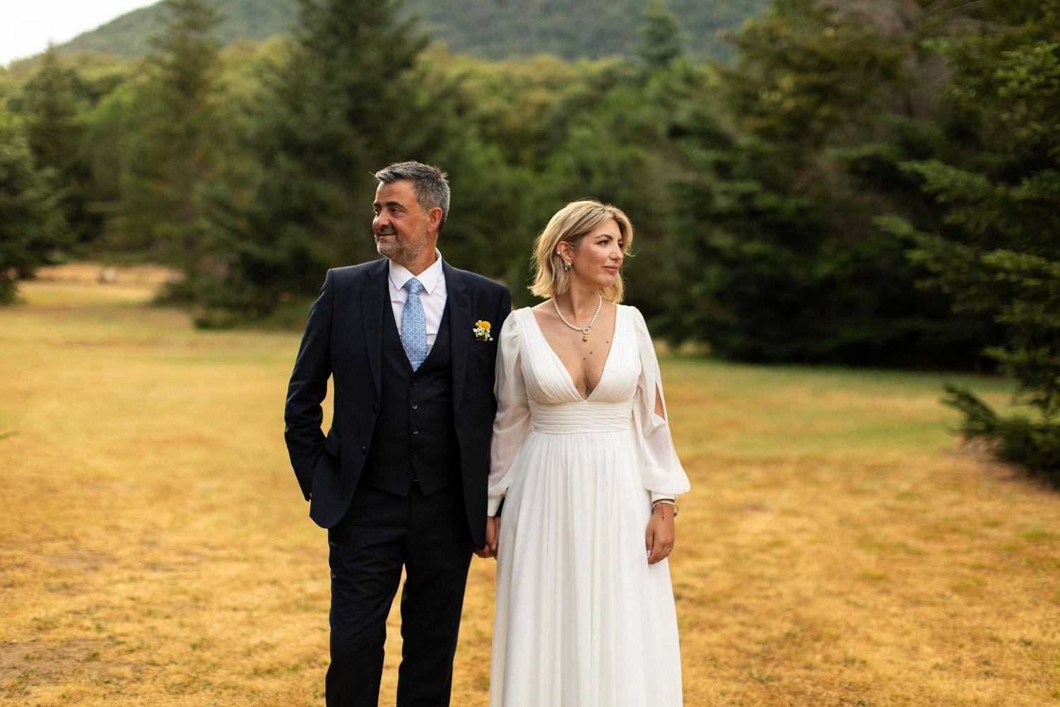 Bride and groom walk hand in hand through golden field with green forested hills.
