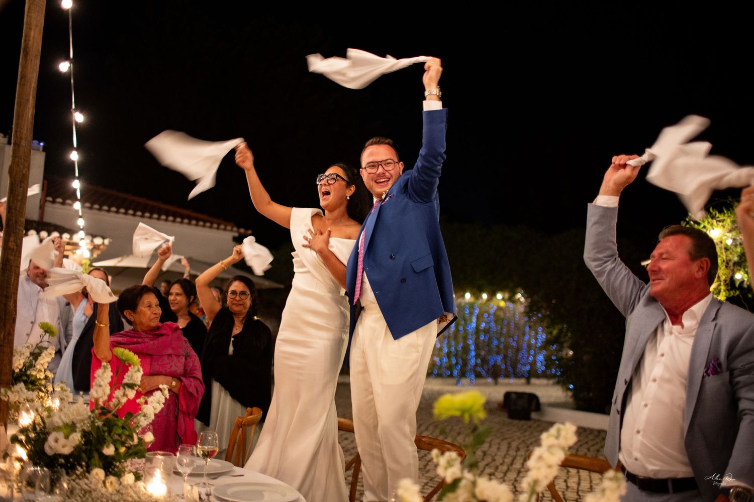 Bride and groom cheer with guests waving white napkins at outdoor night reception.