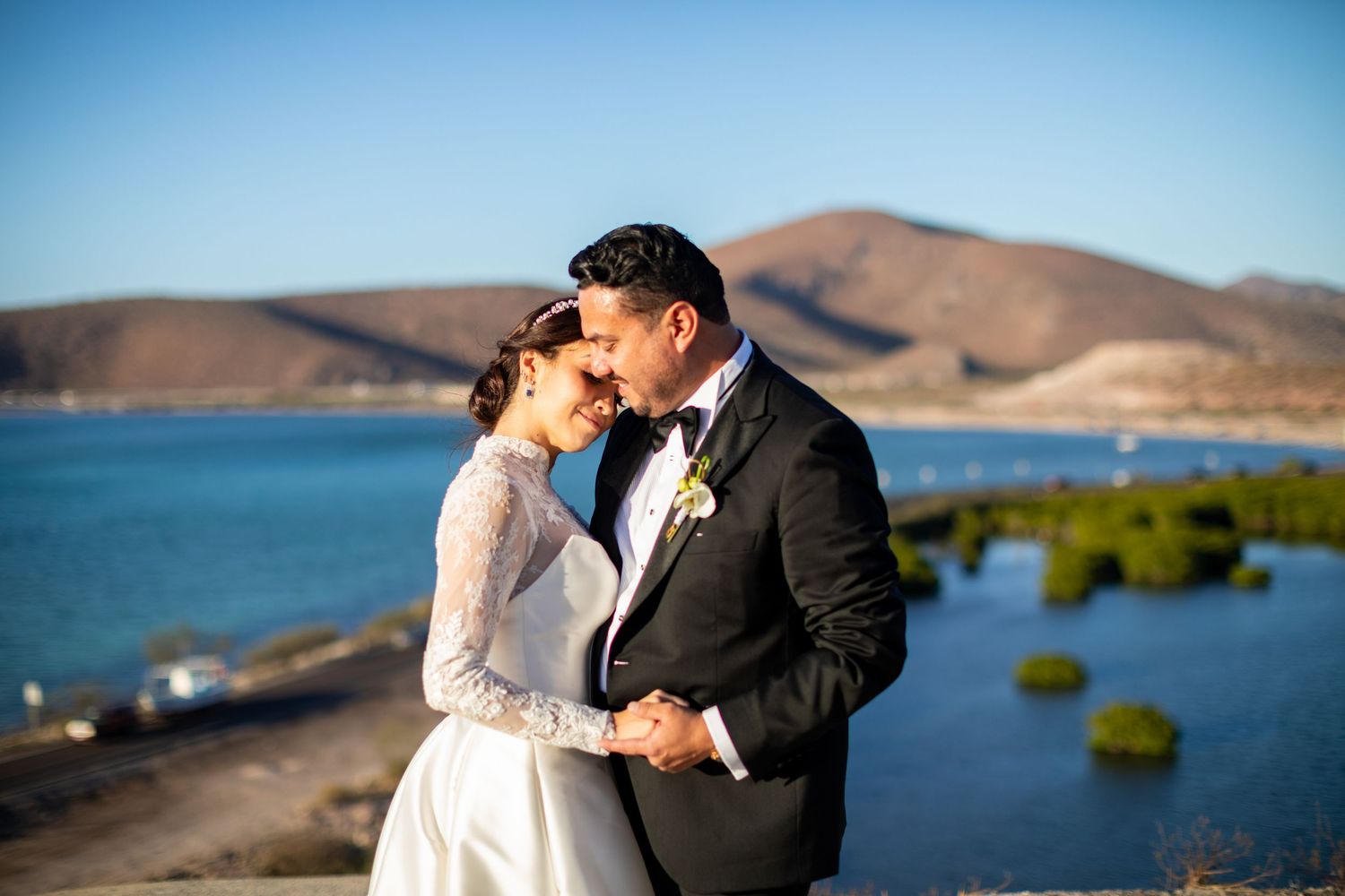 Romantic bride and groom touching foreheads at scenic waterfront location with mountain backdrop.