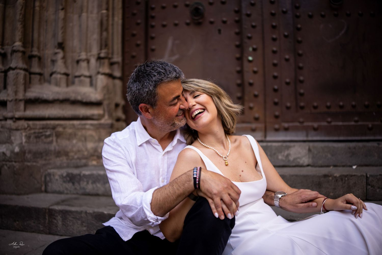 A couple sits on stone steps before an ornate door, sharing a tender moment as the man kisses the laughing woman.