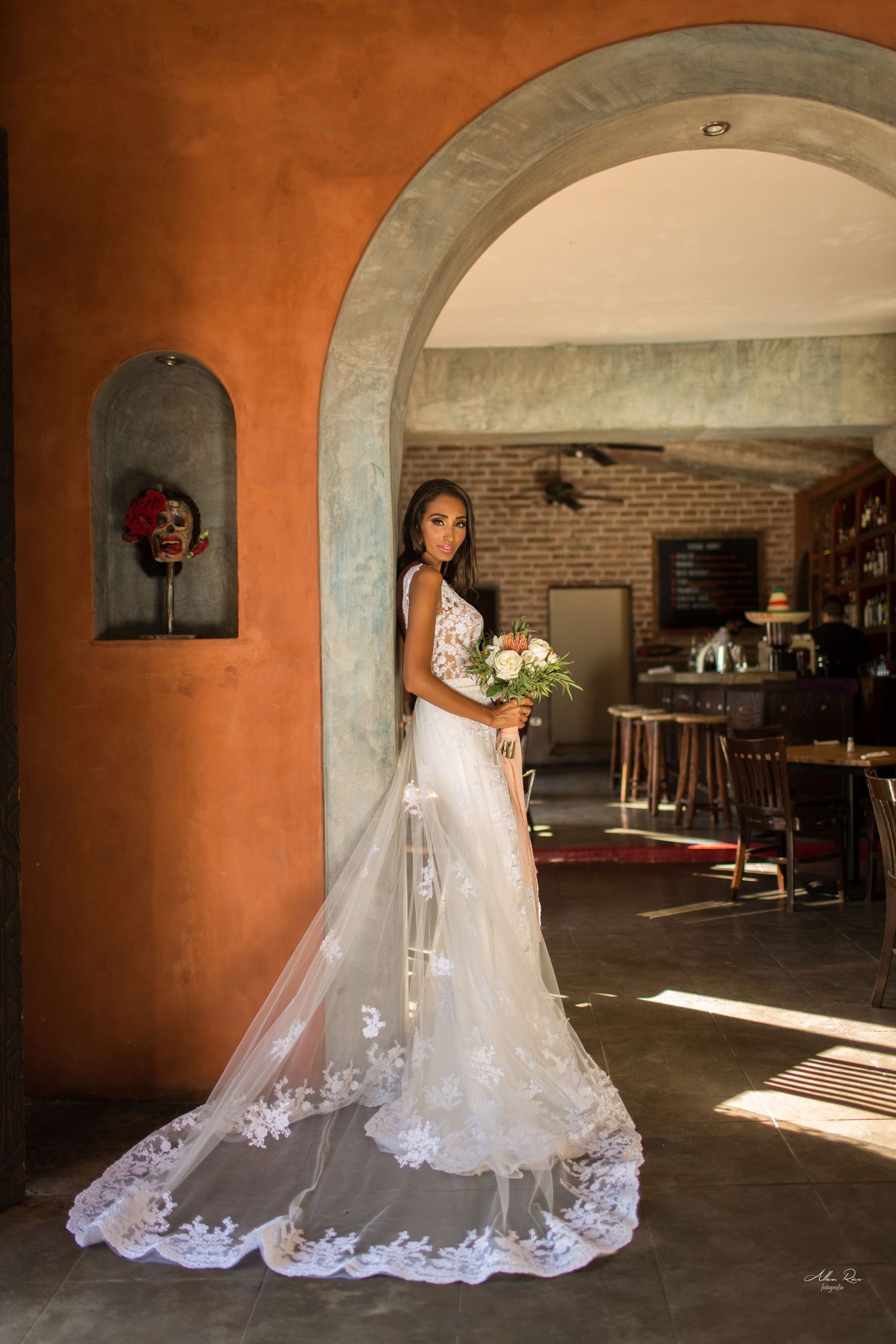 Bride in elegant lace wedding dress with long veil poses under rustic arched doorway in sunlight.
