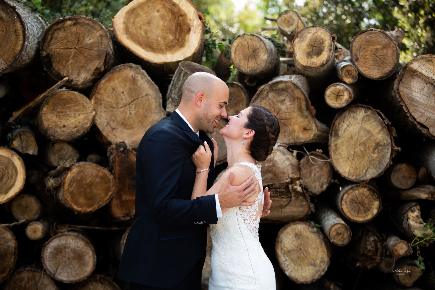 Bride and groom sharing a kiss in front of a rustic stack of cut wooden logs outdoors.