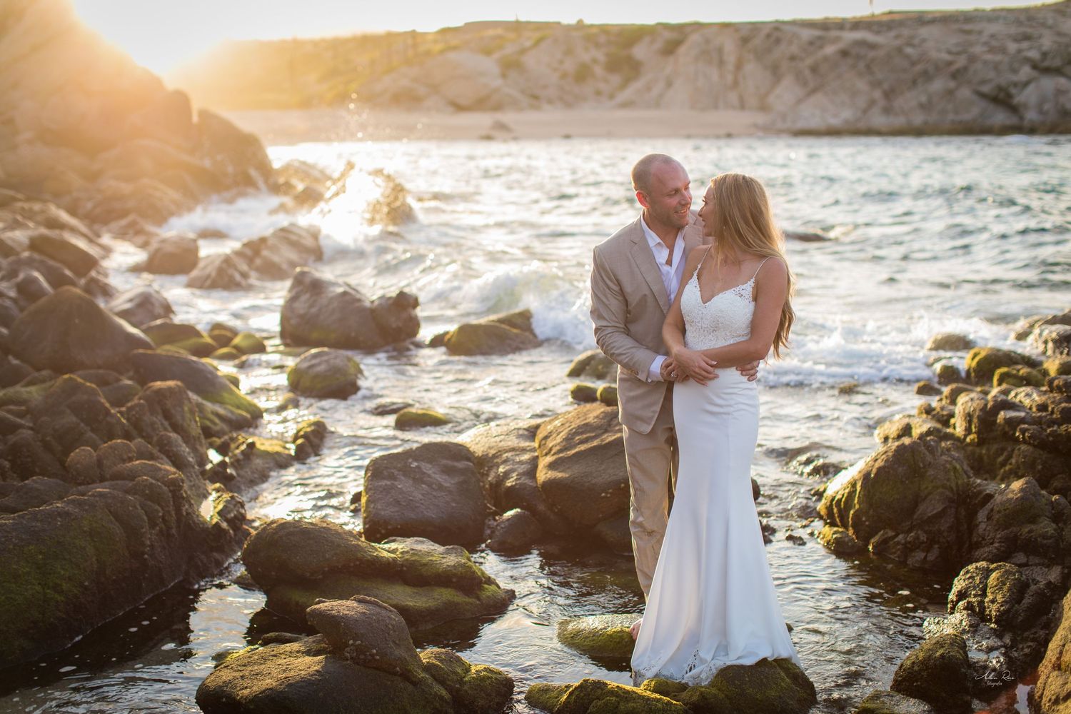 Bride and groom embrace romantically among moss-covered rocks with golden sunset ocean backdrop.