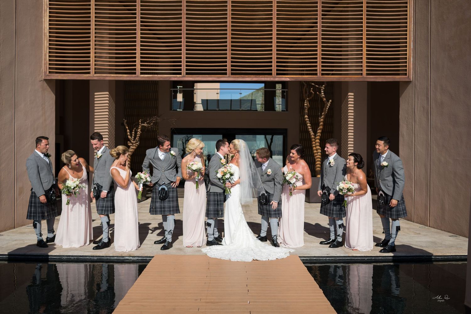 Wedding party with kilted groomsmen and blush bridesmaids celebrate with couple kissing at modern venue.
