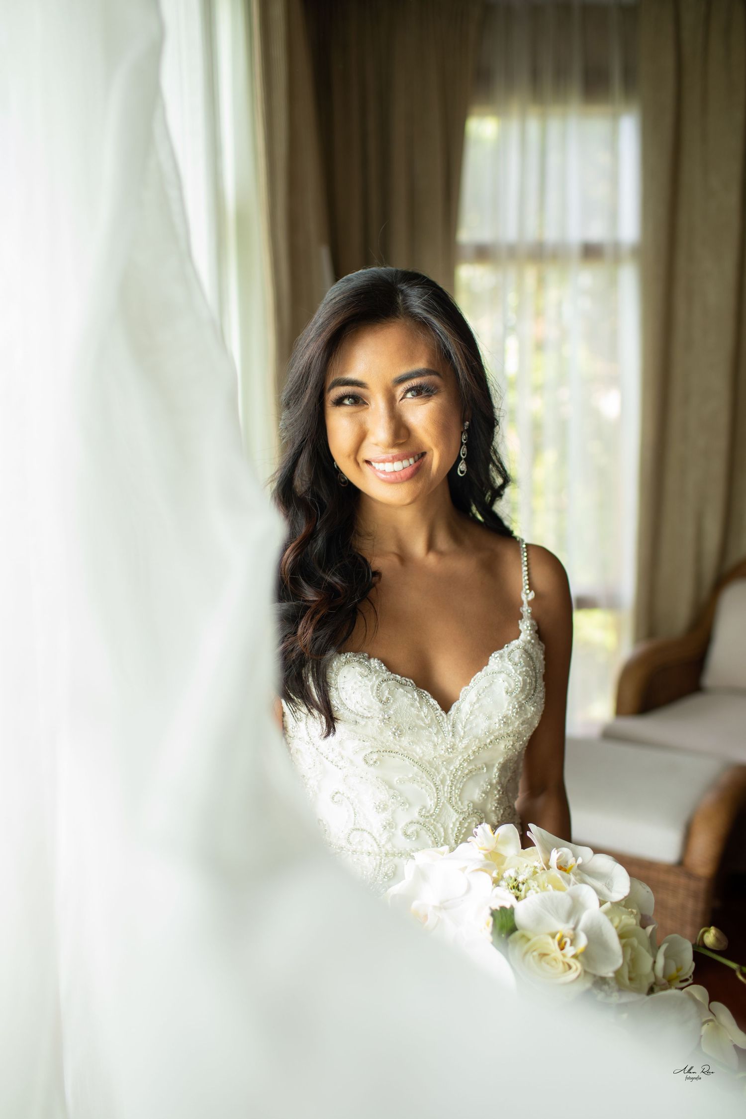 Beautiful smiling bride in beaded lace gown holding white orchid bouquet by window.