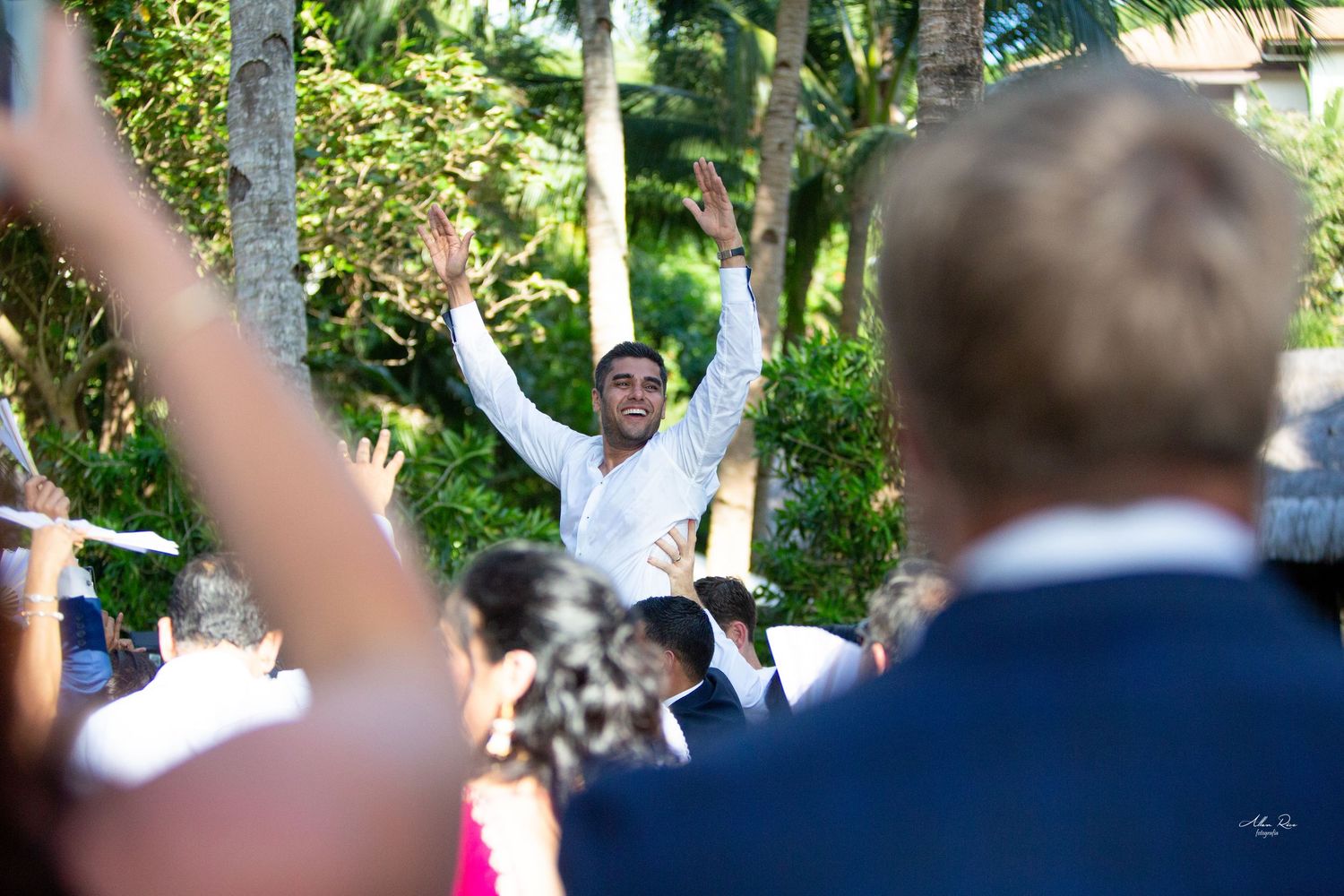 Groom celebrating with raised arms among wedding guests outdoors surrounded by tropical palm trees.