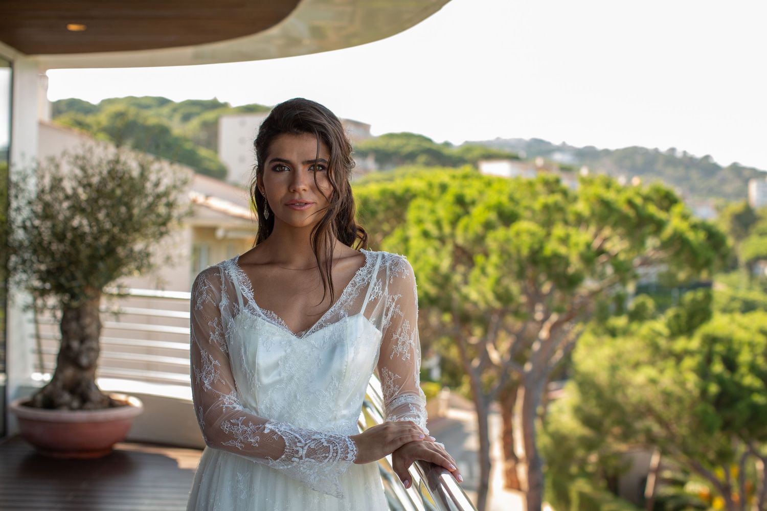 Brunette bride in elegant lace wedding gown stands on a balcony surrounded by lush green trees.
