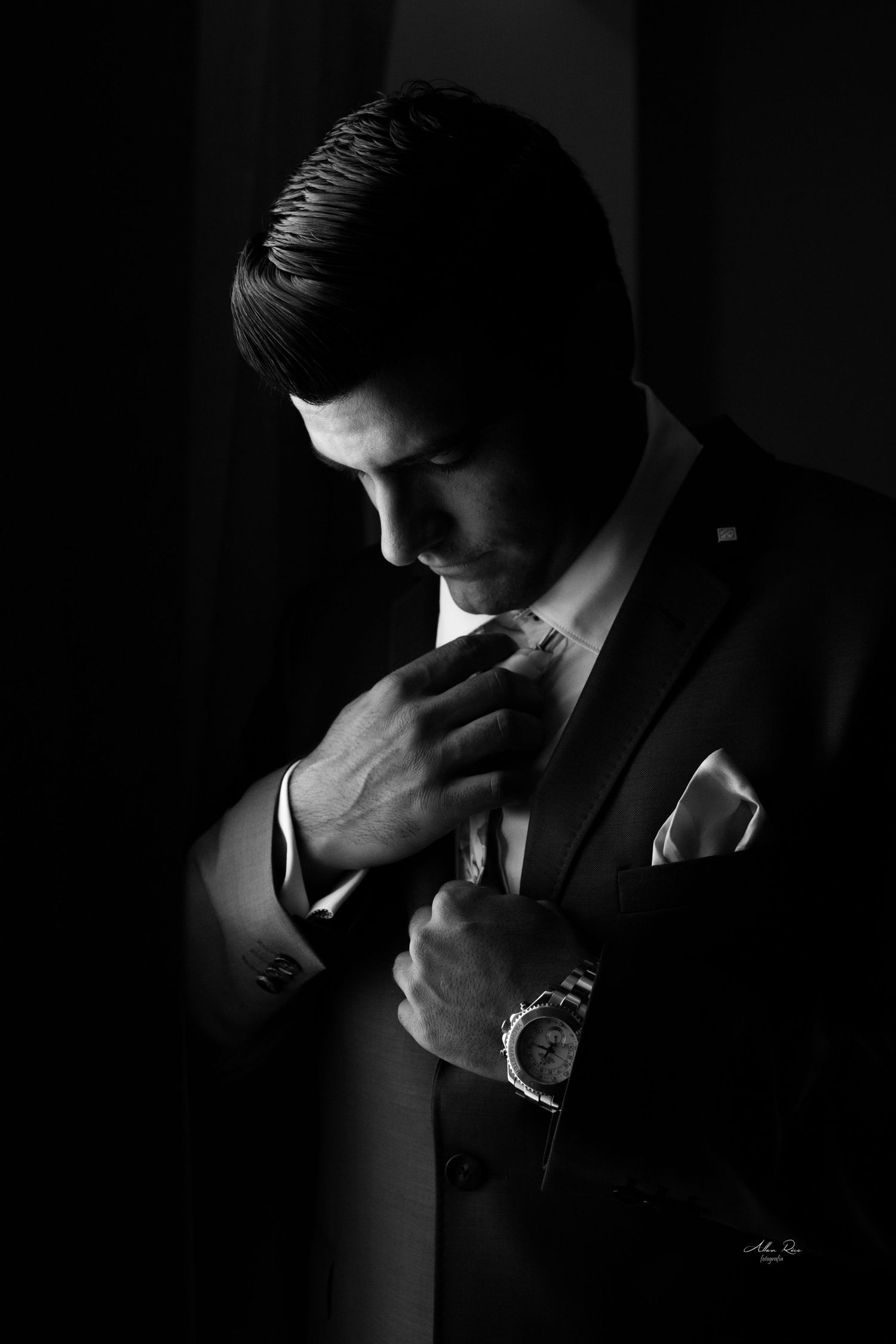 Dramatic black and white portrait of groom in dark suit adjusting tie with luxury watch on wrist.