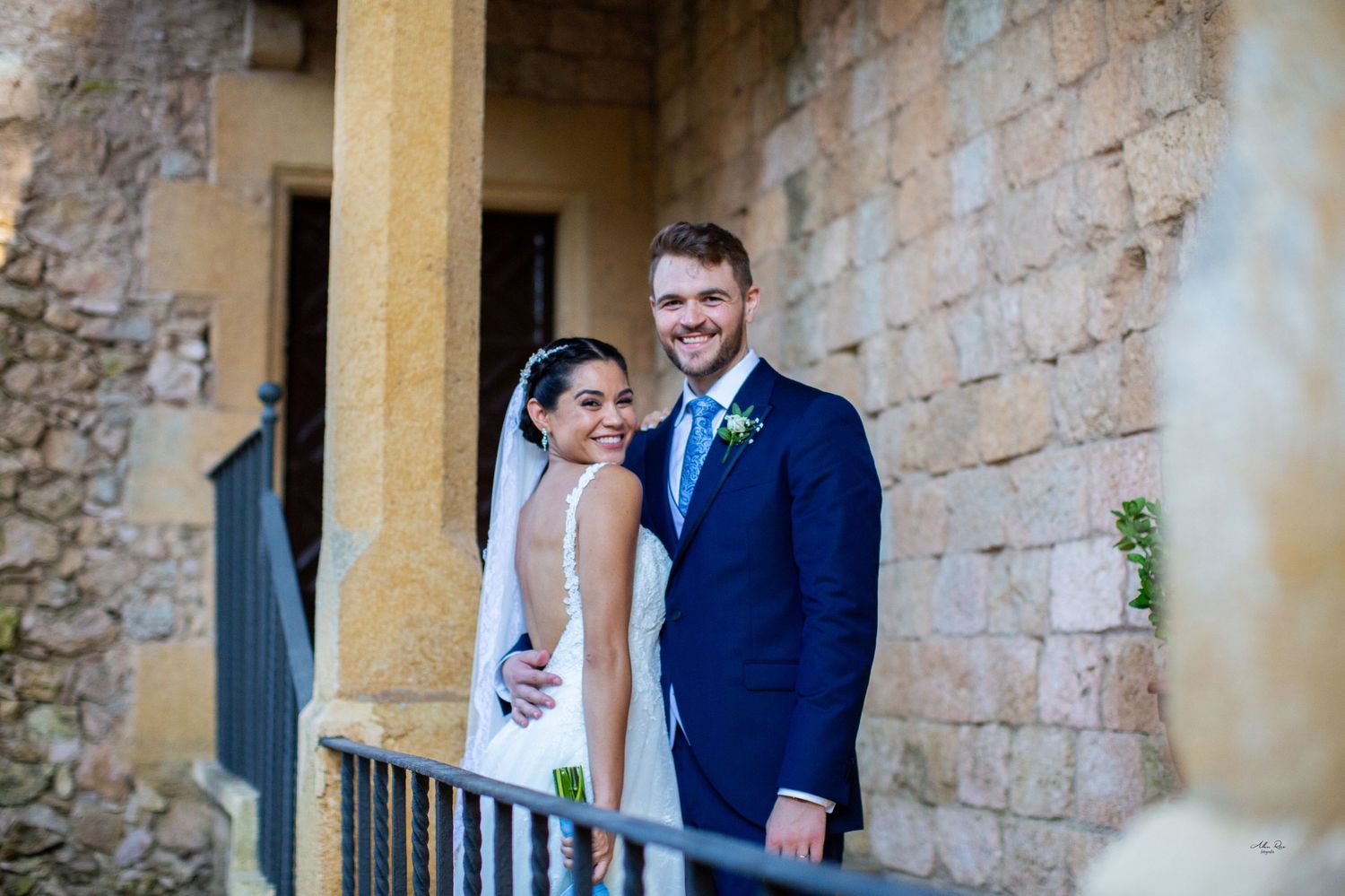 Smiling bride and groom posing together against rustic stone wall during outdoor wedding portrait session.