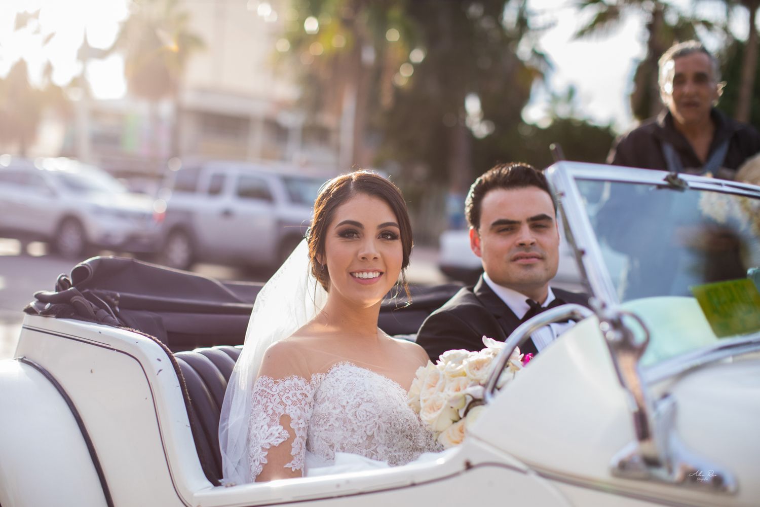 Bride and groom riding in vintage white convertible car after outdoor wedding ceremony at sunset.