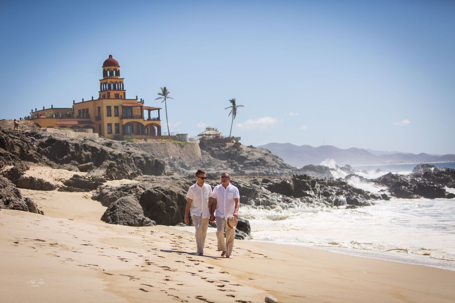 Two grooms in casual attire walk hand in hand along a sunny beach.
