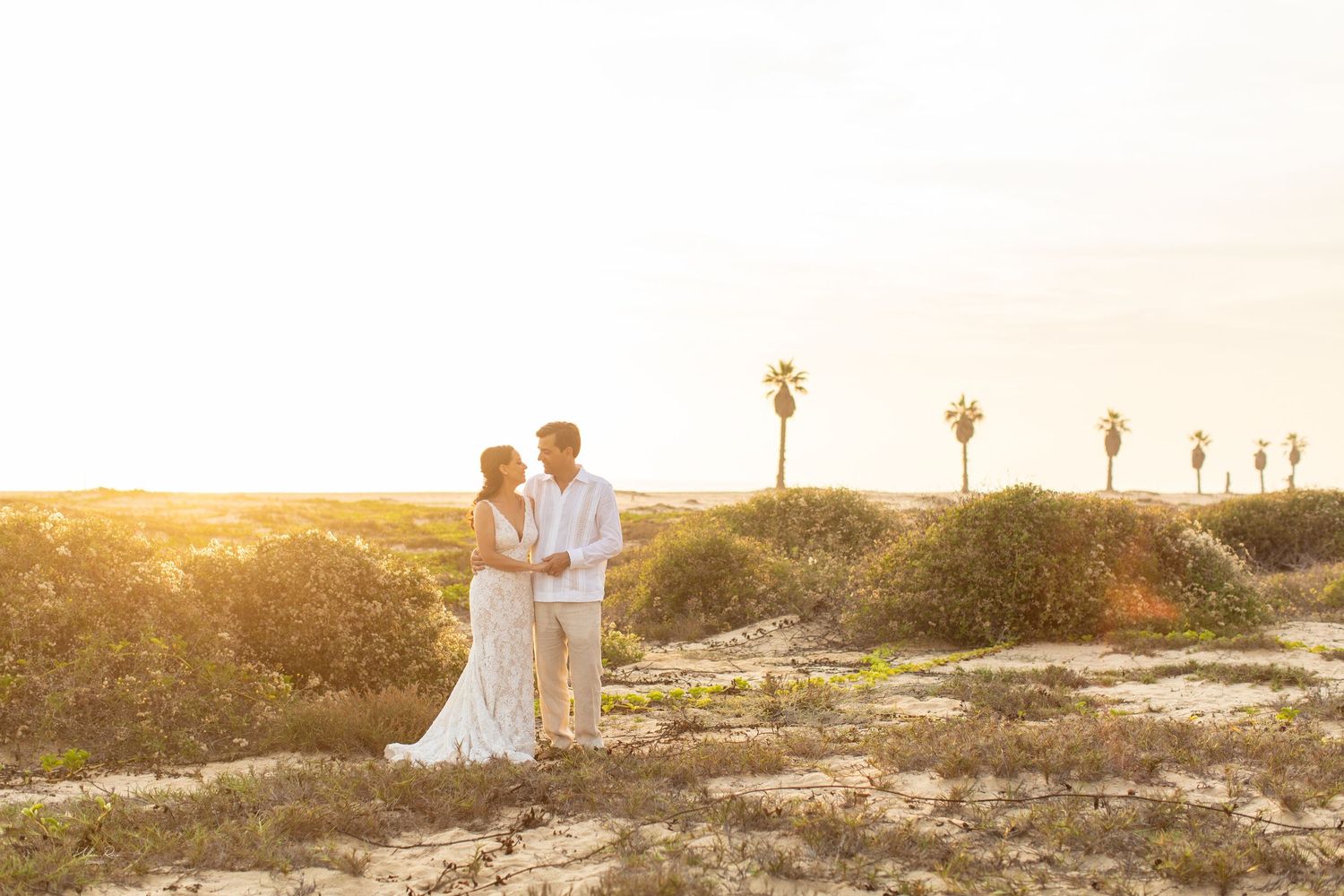 Bride and groom standing together in golden sunset light among desert shrubs with palm trees.