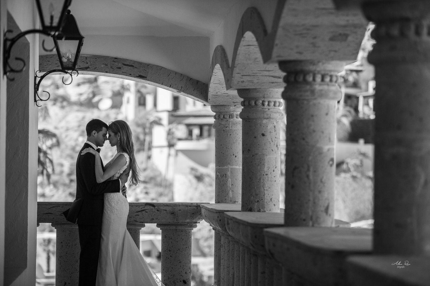 Bride and groom share tender embrace between ornate stone columns on a classic architectural balcony.