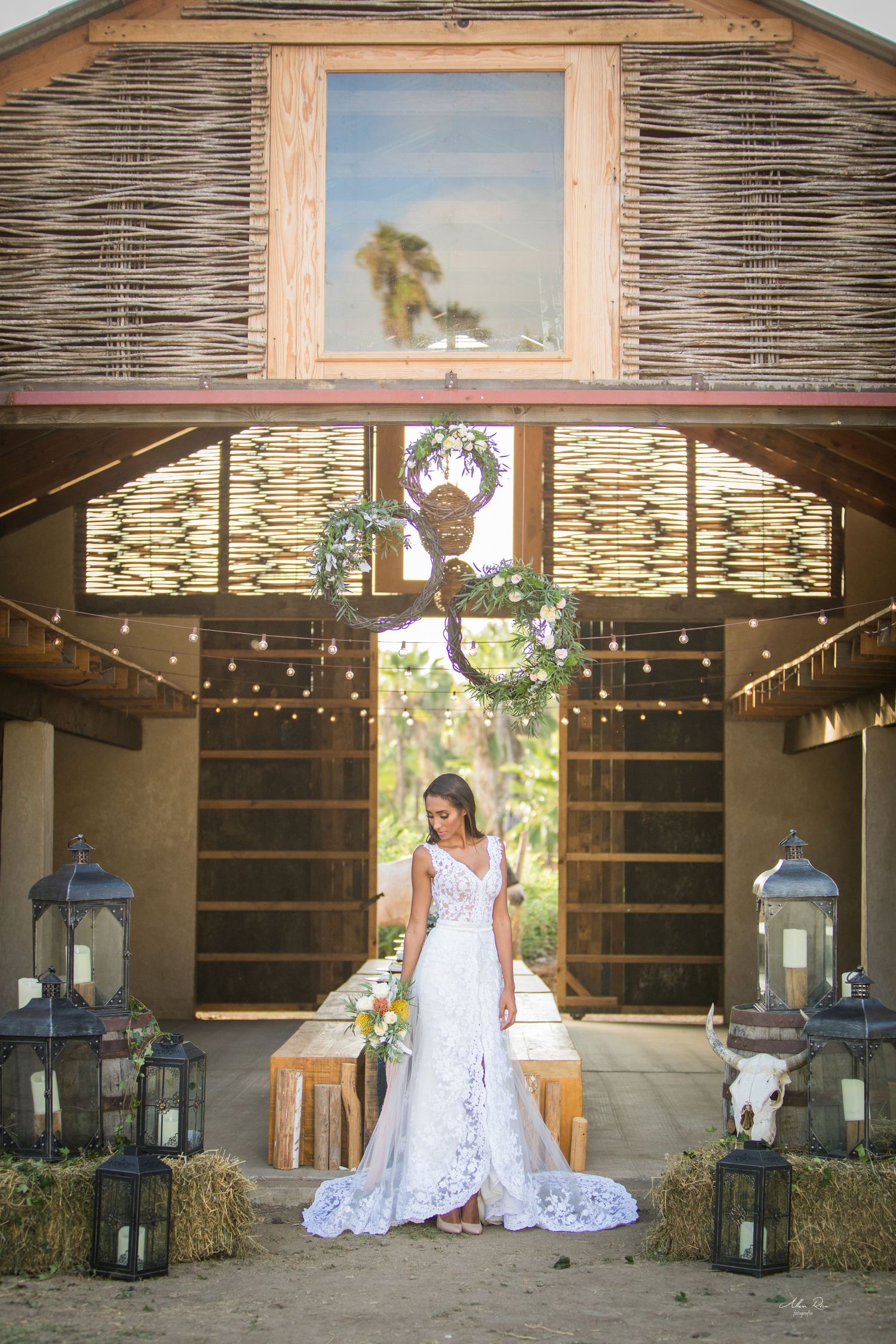 Bride in lace gown stands before rustic barn altar with hay bales and lanterns at outdoor wedding.