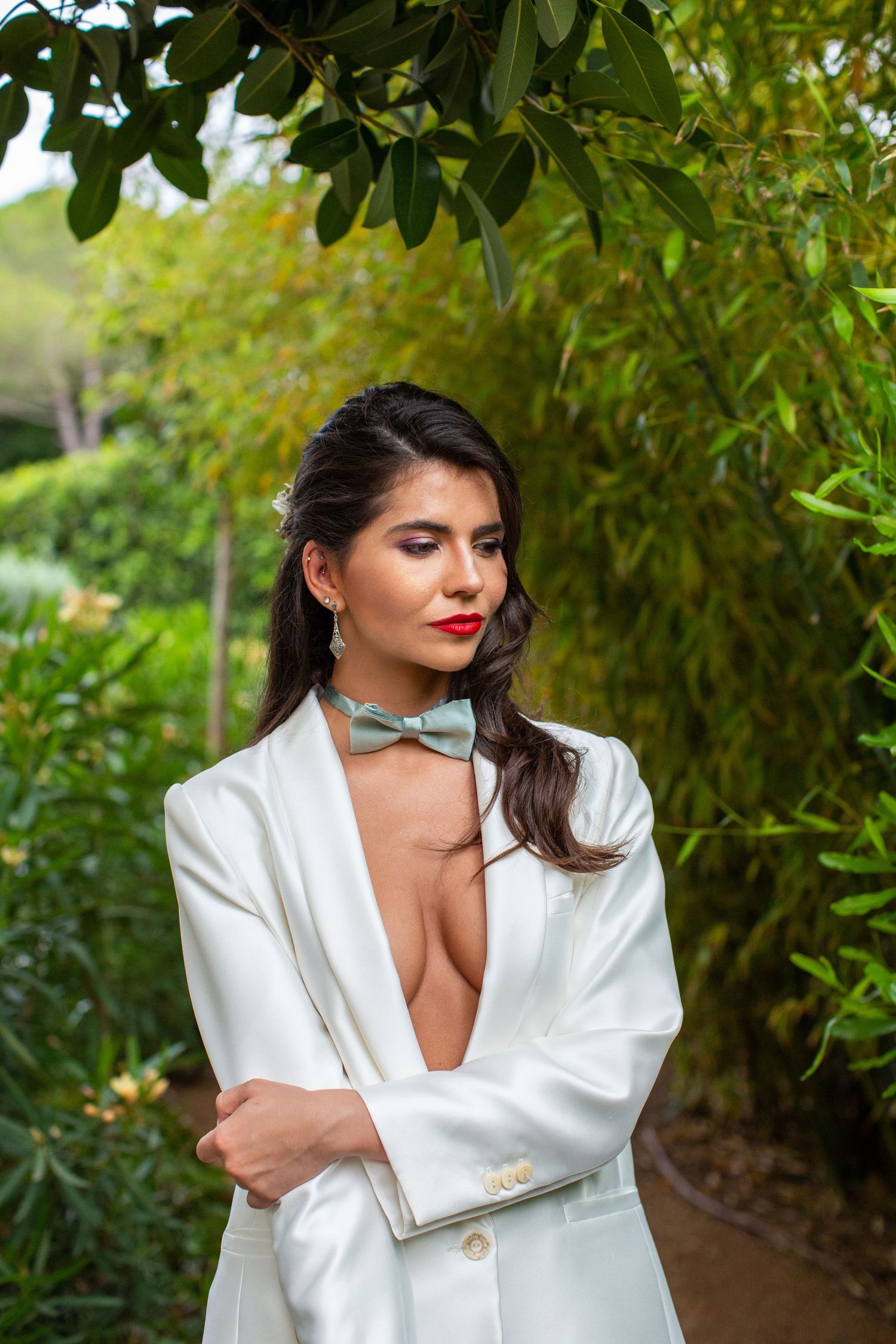 Elegant bride in a plunging white blazer suit with sage bow tie stands thoughtfully among lush bamboo greenery.