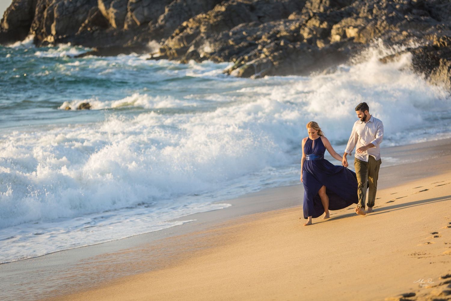 Couple walking hand in hand on a sandy beach with dramatic crashing waves at golden hour.
