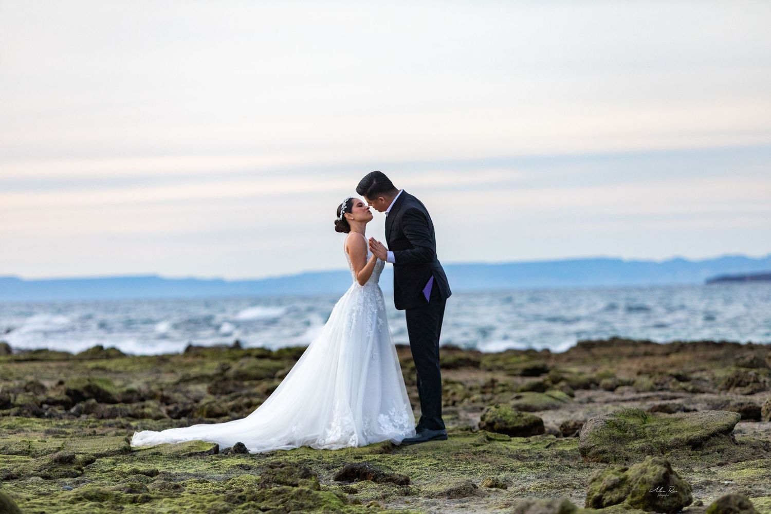 Bride and groom kissing on moss-covered rocks with ocean waves in the background.