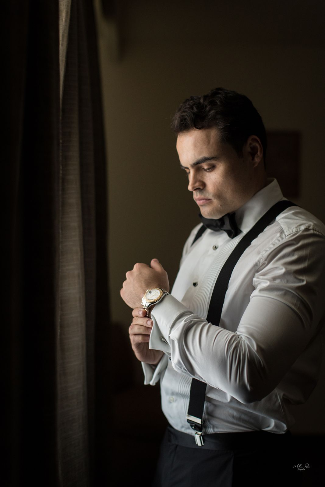 Groom in suspenders and bow tie adjusting his gold watch near a dark curtain.