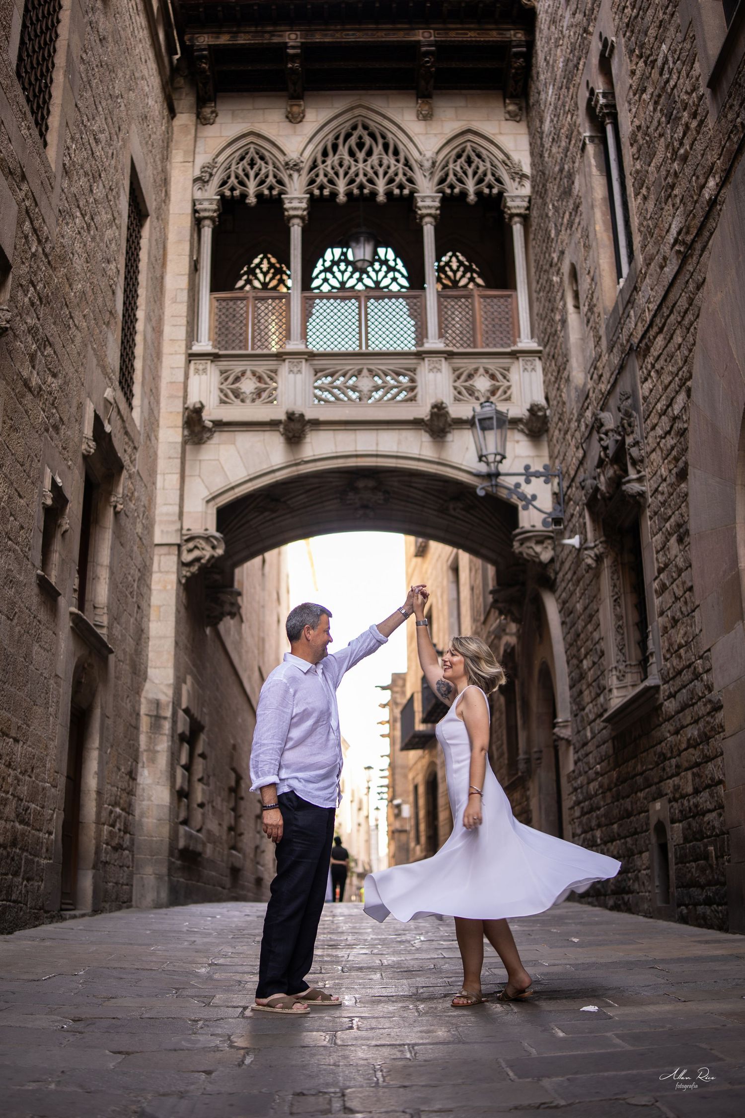 A couple dances in a narrow Gothic Quarter alley beneath an ornate medieval stone bridge in Barcelona.