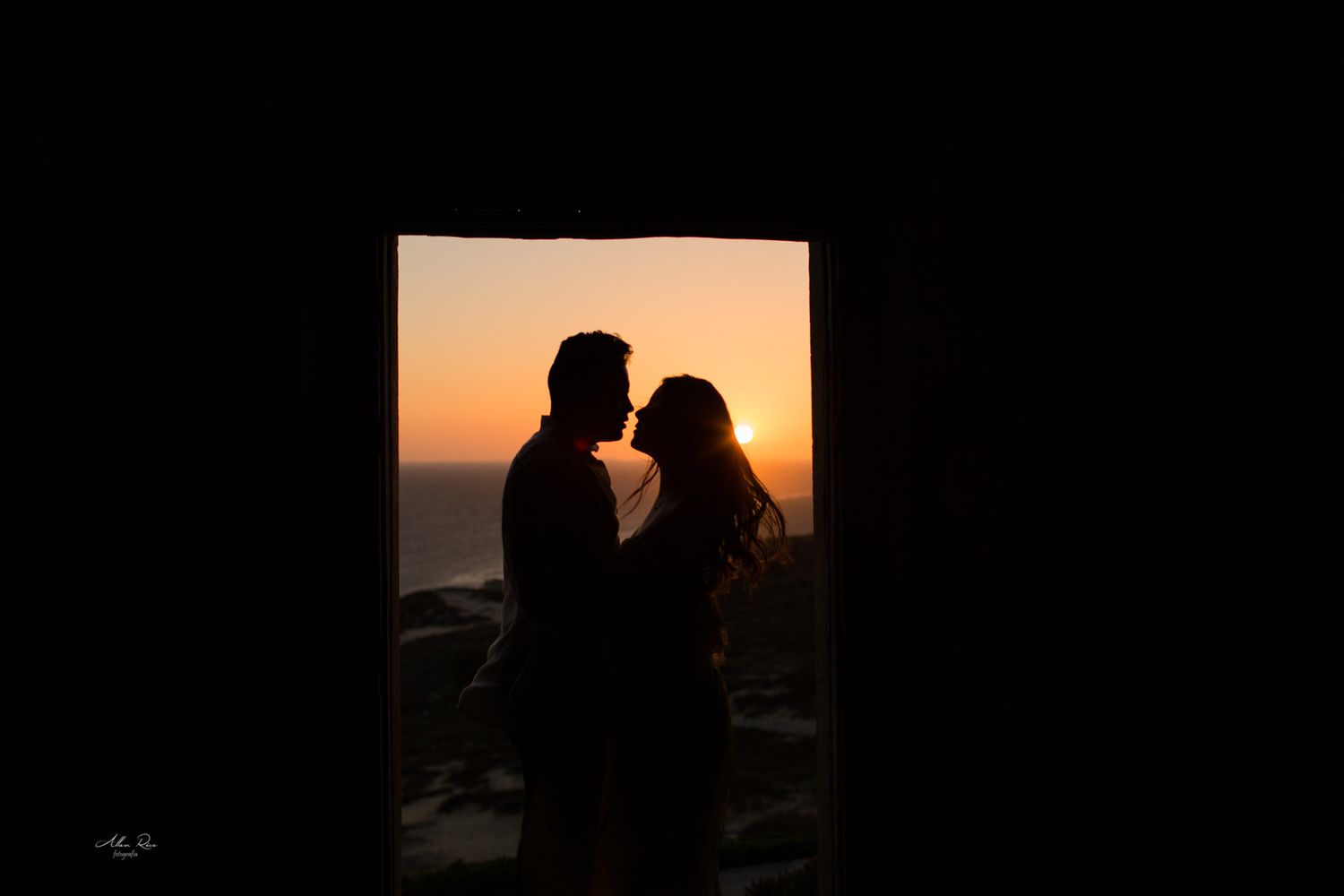 Romantic couple silhouette sharing a kiss framed by a dark doorway at sunset.