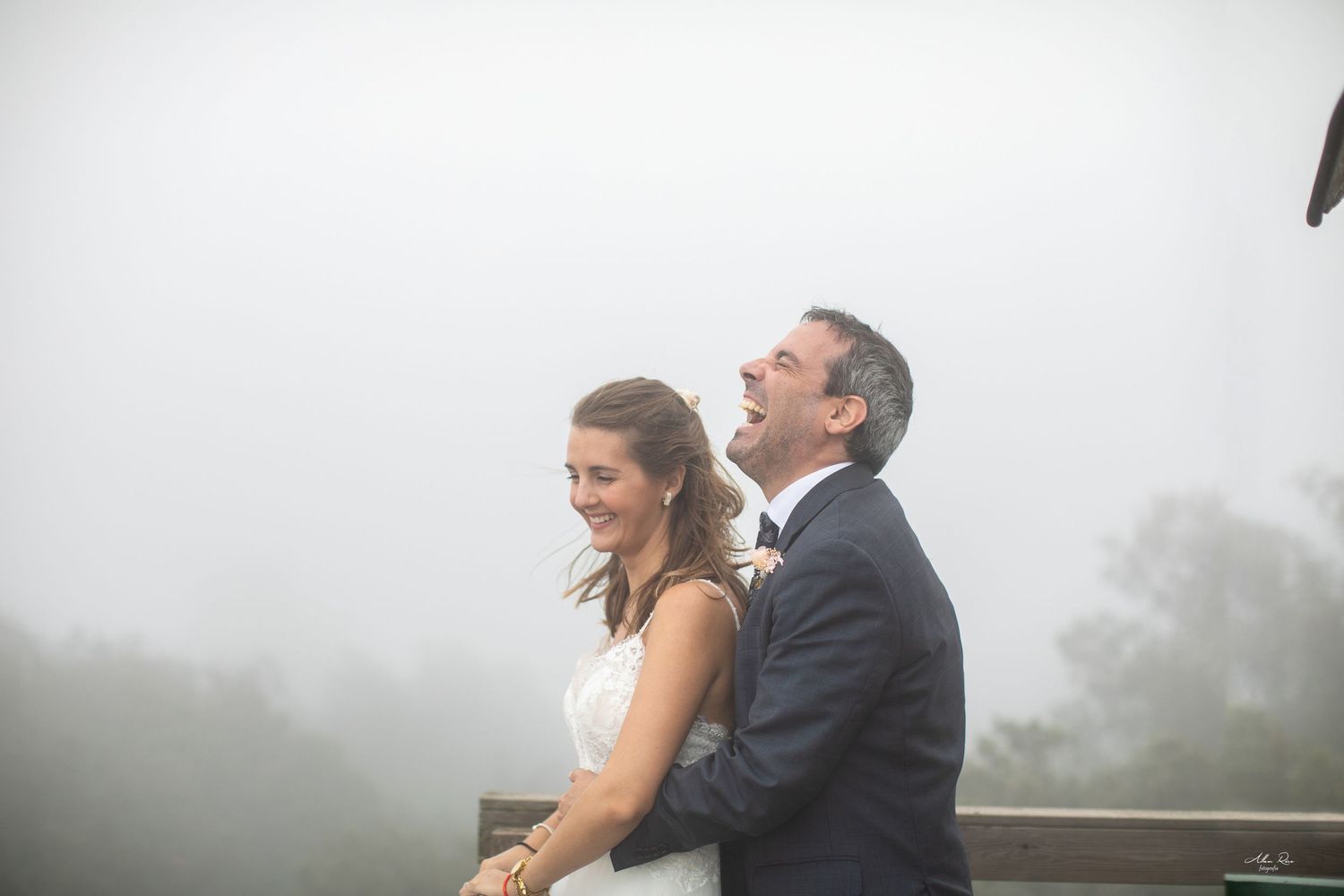 Laughing bride and groom pose together on misty mountain overlook on their wedding day.