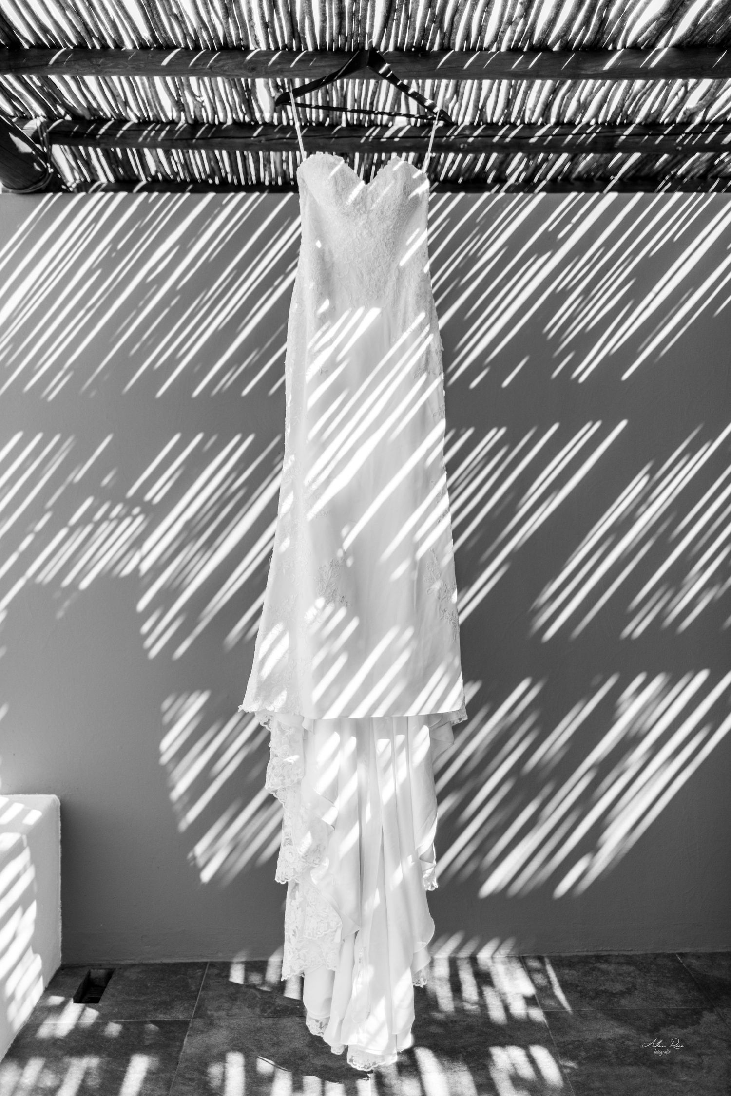 Black and white photo of a strapless wedding gown hanging under a pergola casting dramatic diagonal shadows.