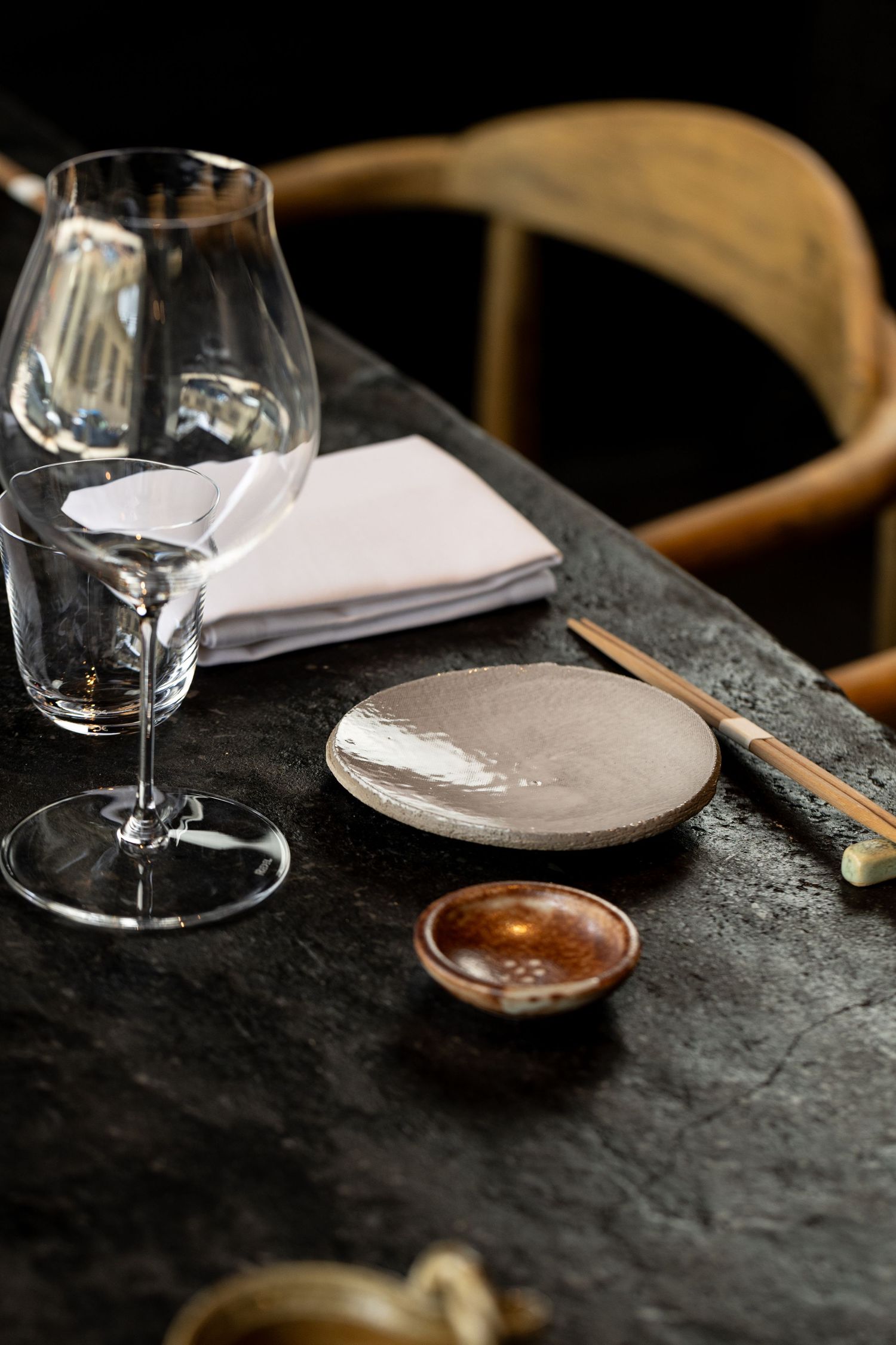 Elegant Japanese table setting with crystal wine glass, ceramic plate, chopsticks, and small bowl on dark stone surface.