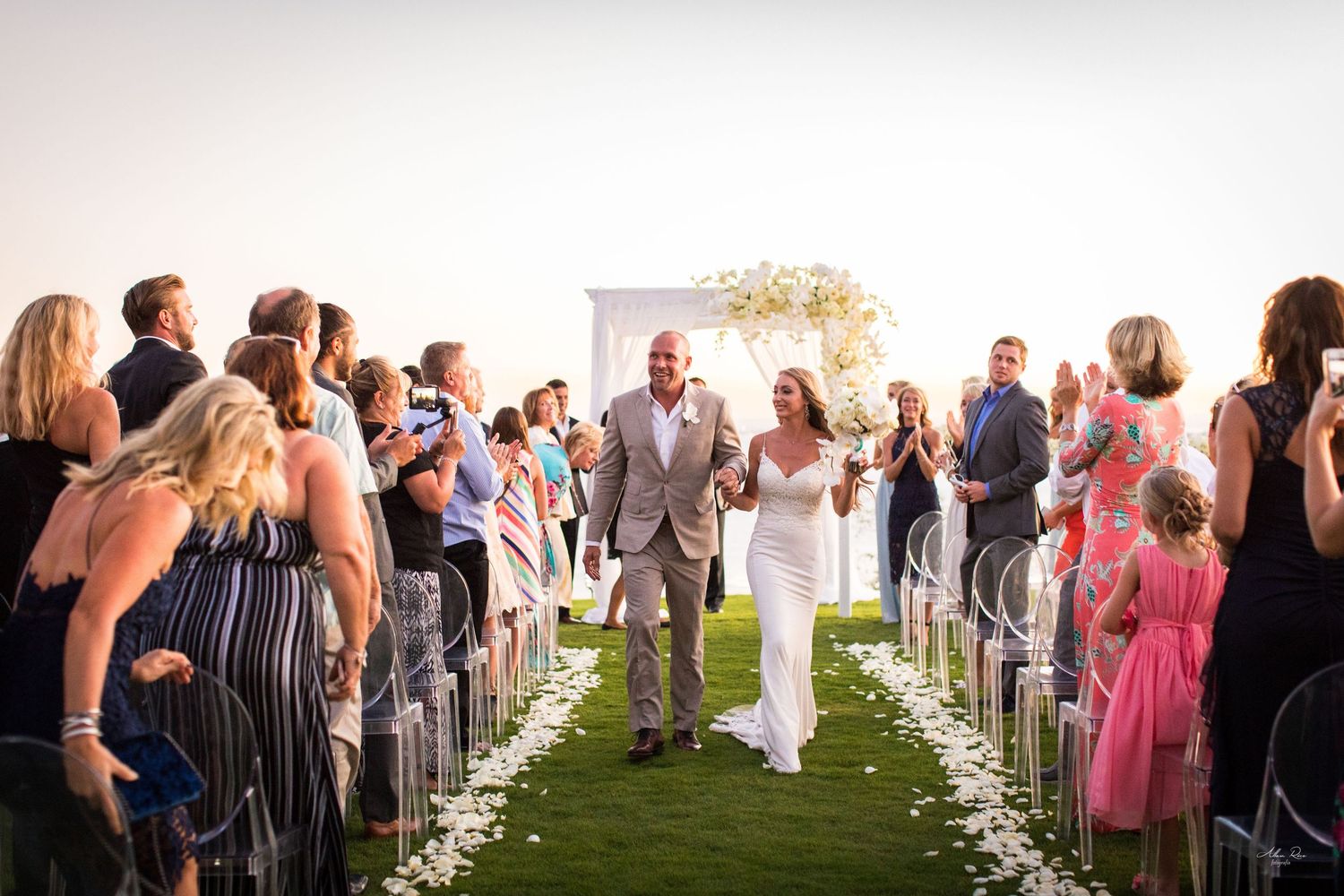 Joyful newlyweds walk down petal-lined aisle as guests celebrate at stunning outdoor oceanfront ceremony.