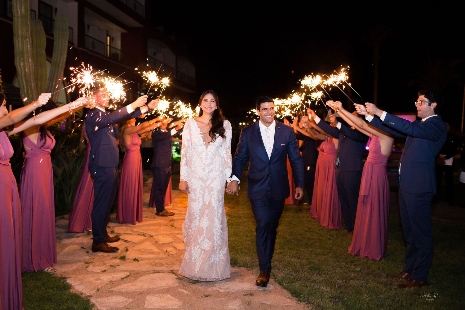 Bride and groom walk through sparkler tunnel with wedding party in pink dresses at night celebration.