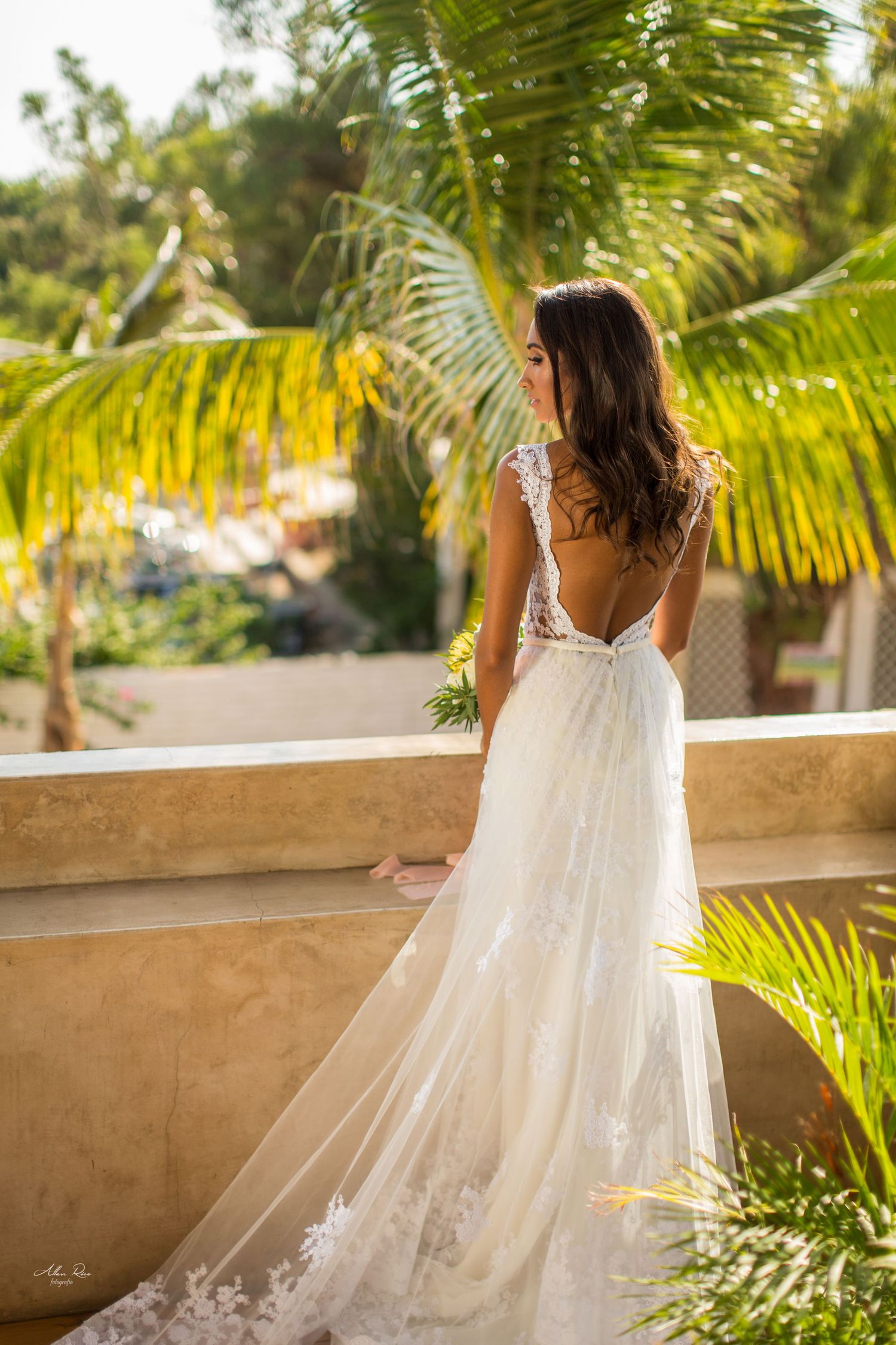 Bride in backless lace wedding dress with long train stands on balcony surrounded by palm trees.