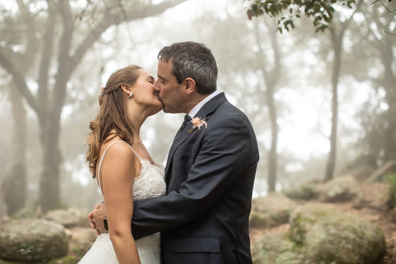 Newlyweds share a romantic kiss in a foggy woodland setting on their wedding day.