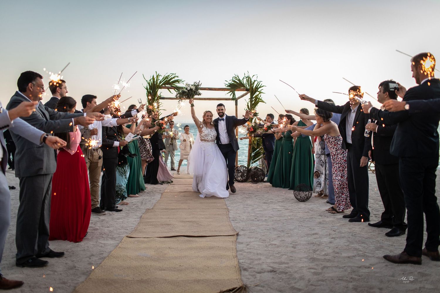 Bride and groom celebrate sparkler exit surrounded by cheering guests at romantic beach wedding.