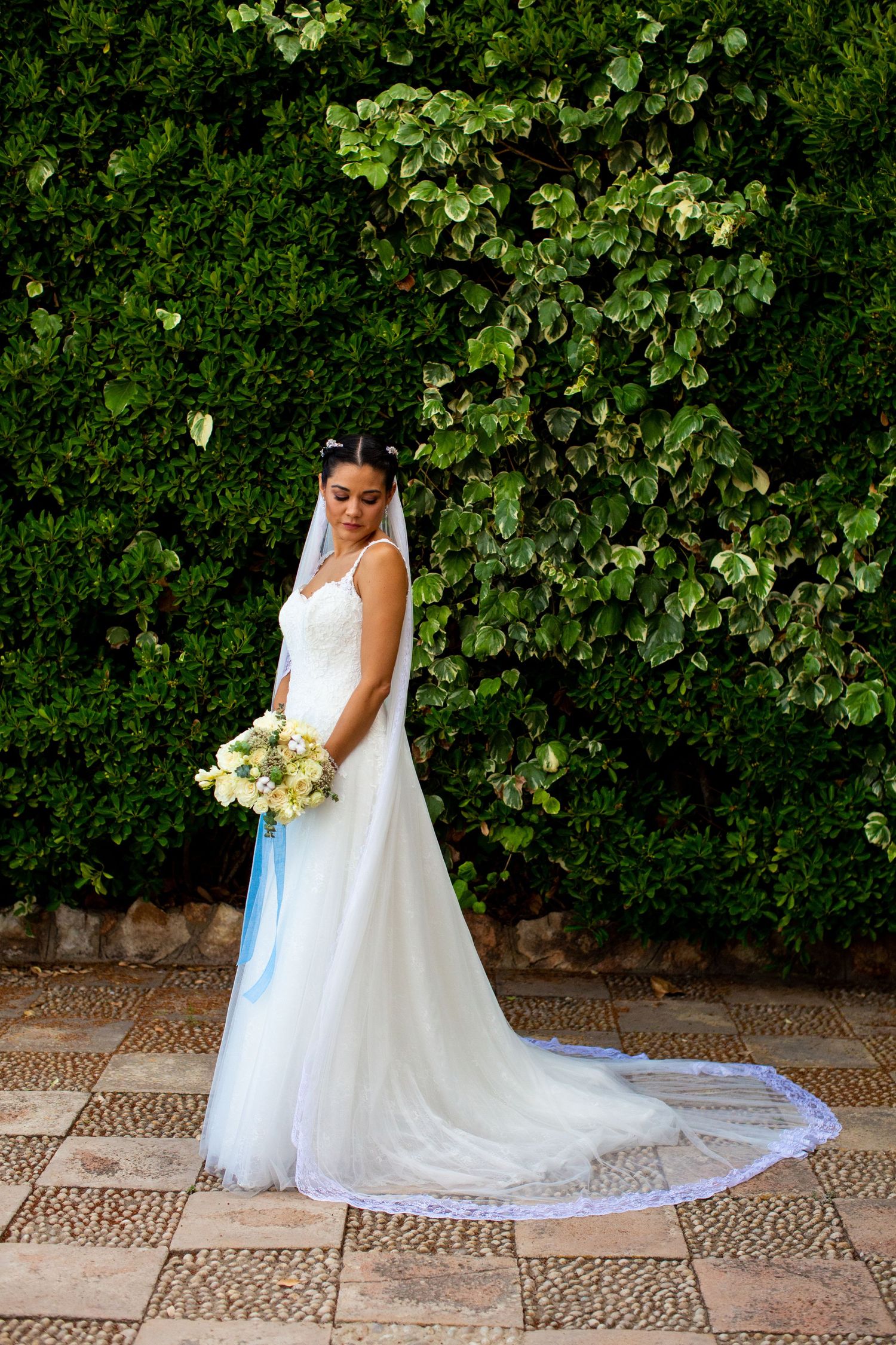 Bride in white gown and veil holding a bouquet poses before a lush green ivy wall outdoors.