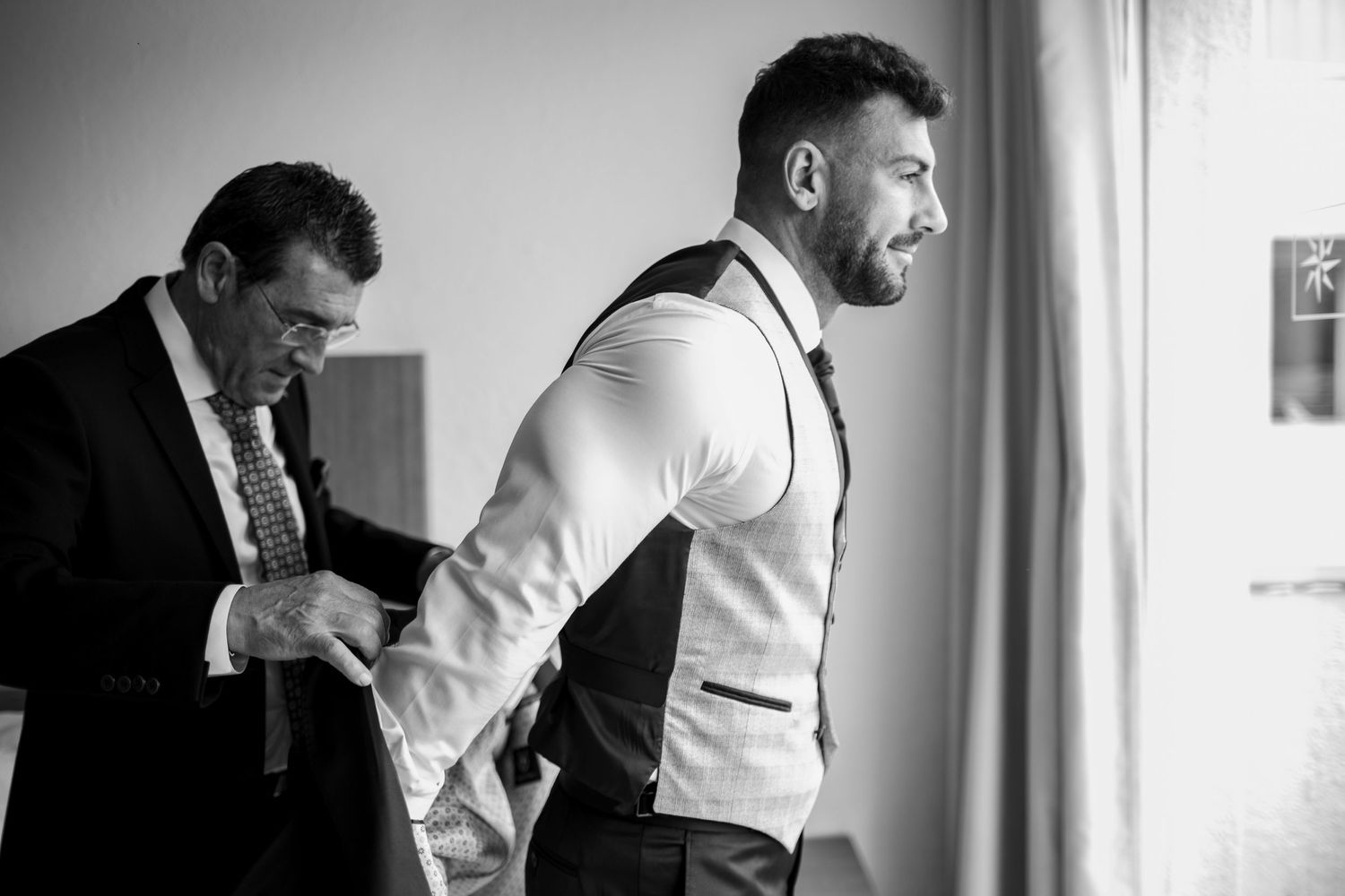 Black and white photo of groom being helped into vest by older man before wedding ceremony.