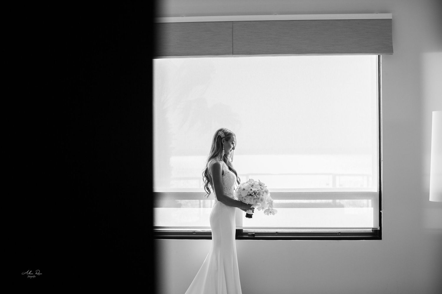 Elegant bride in wedding gown holding bouquet silhouetted against large window in black and white photo.